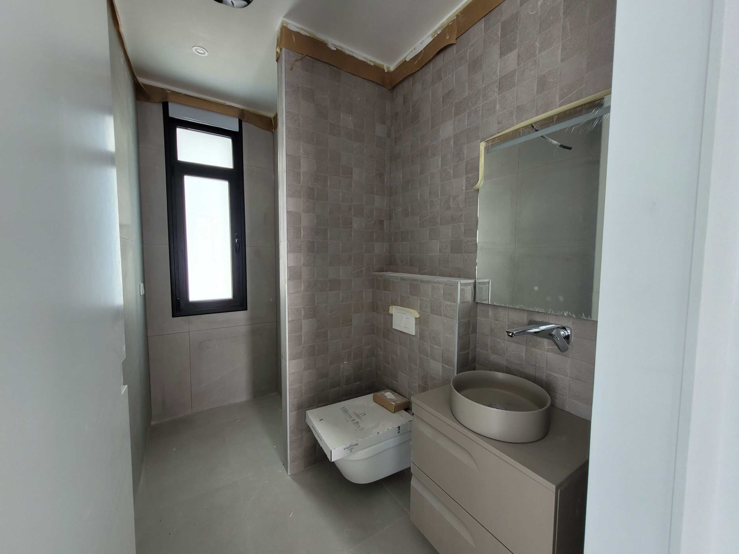 Modern badkamer met beige tegelwand, wandcloset, ronde wastafel en zwart raam voor natuurlijk licht – Costa Concepts.